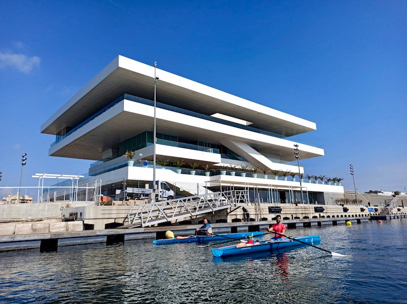 Foto remando en el bote de remo de mar individual y al lado Nacho con oro bote, detrás el edificio Veles e Vents de Valencia